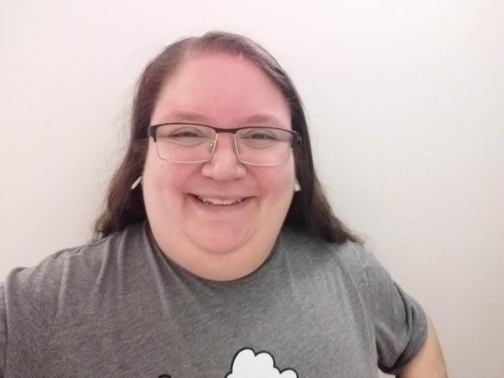 A person with long brown hair and glasses stands in front of a white background, smiling.