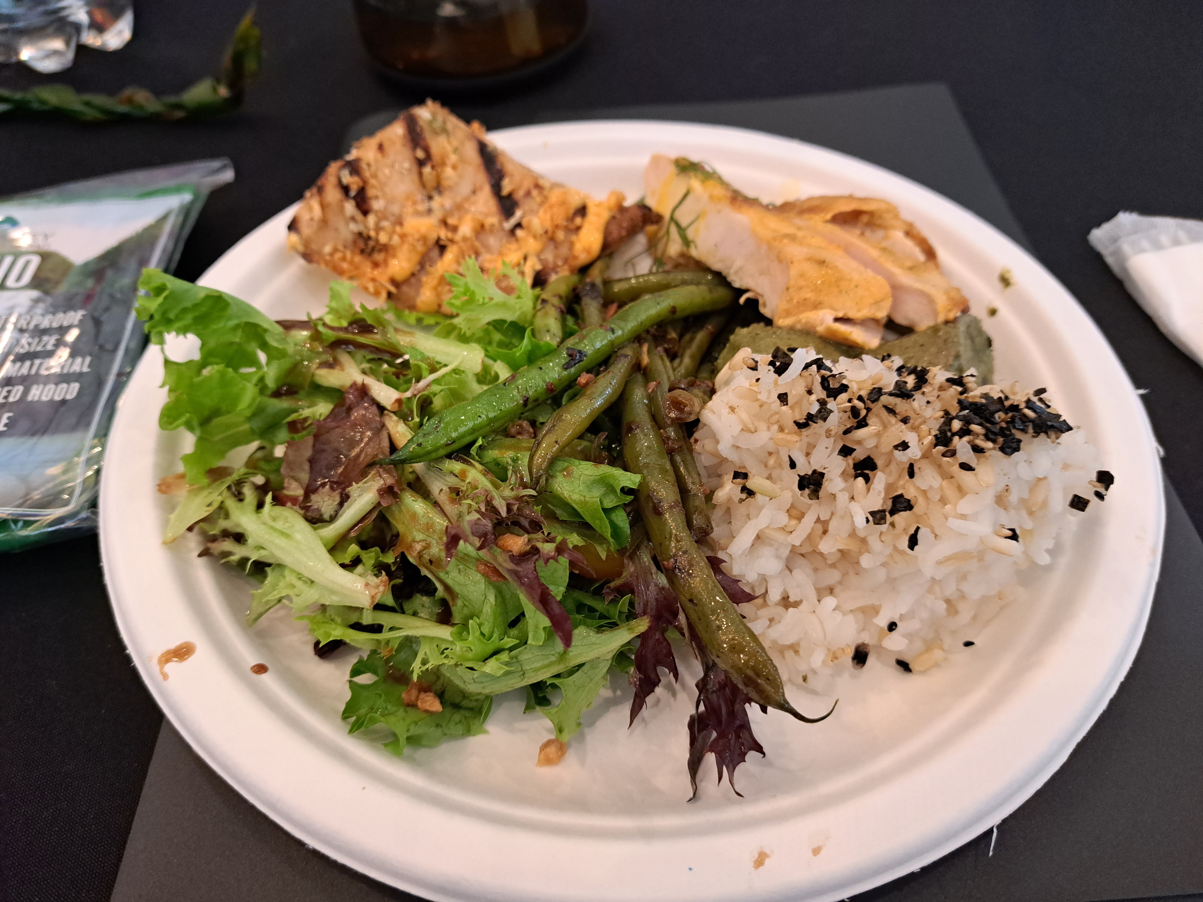 A close-up image of a paper plate full of food on a black tablecloth. There is a piece of grilled white fish with a spicy mayo drizzle, slices of chicken breast, roasted purple sweet potatoes, white rice with furikake, sesame green beans, and a light salad.
