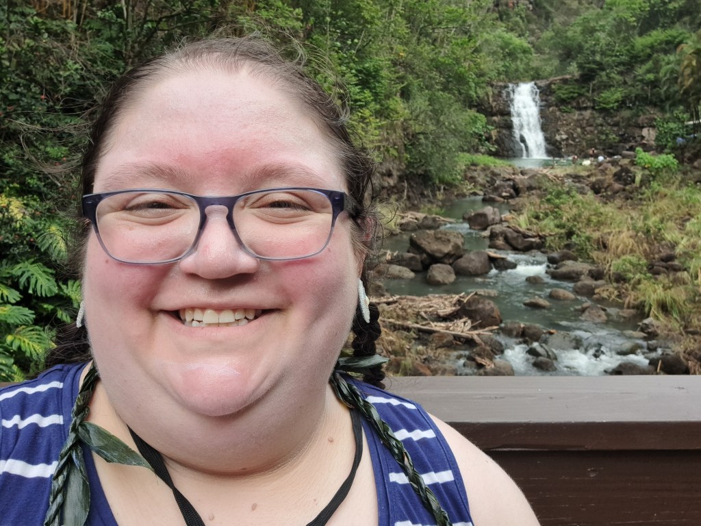 Allie stands smiling in front of a waterfall. The waterfall is distant but still clearly visible. It flows into a pool that then flows into a stream that goes under the bridge Allie is standing on. Allie is wearing a blue tanktop with white stripes, a dark green tea leaf lei, blue glasses, and white beaded earrings. Allie's hair is pulled back into two braids and they are grinning widely.