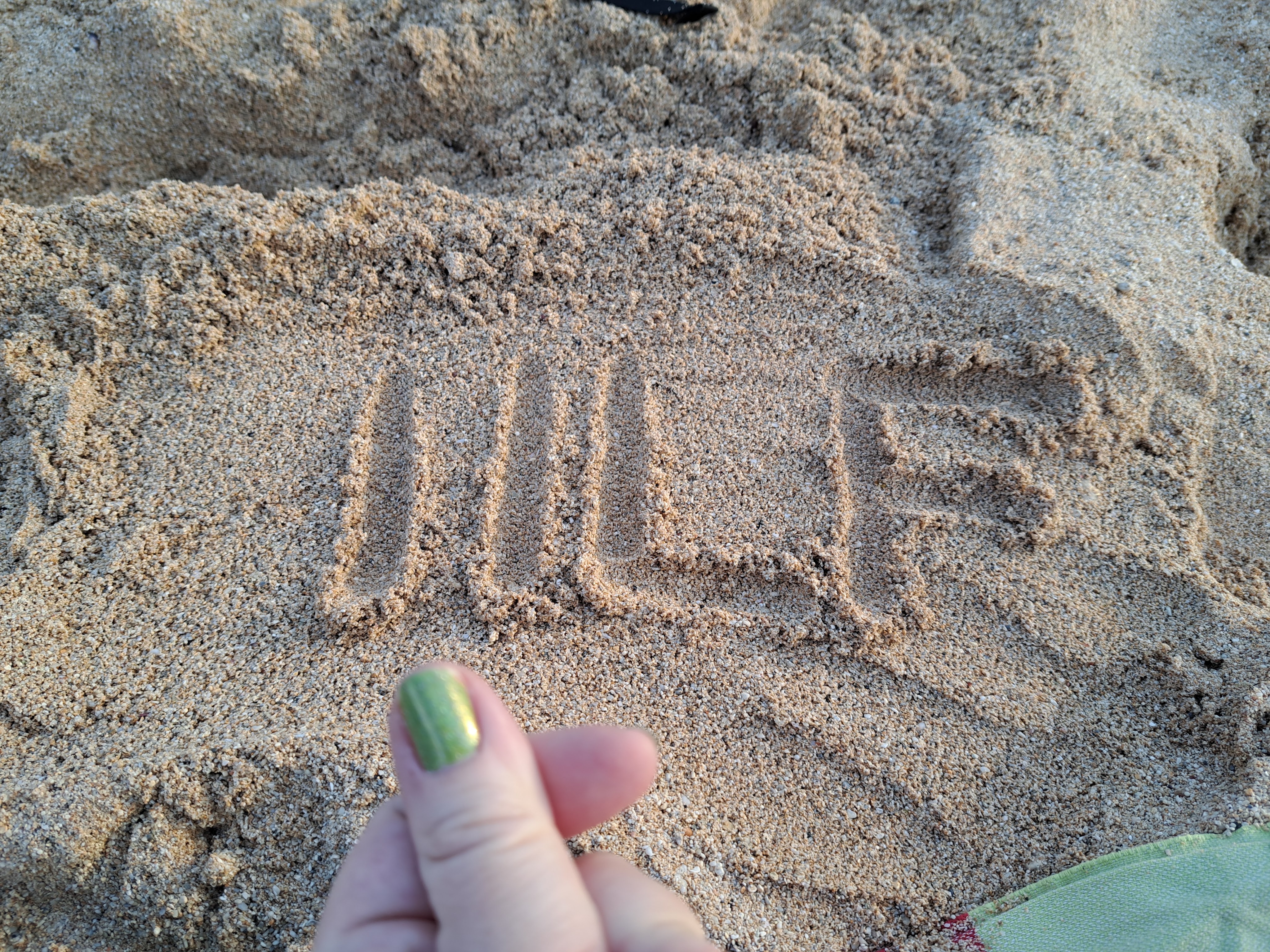 The letters I I L F are traced into beach sand. Allie's thumb and forefinger are crossed, making a small heart.